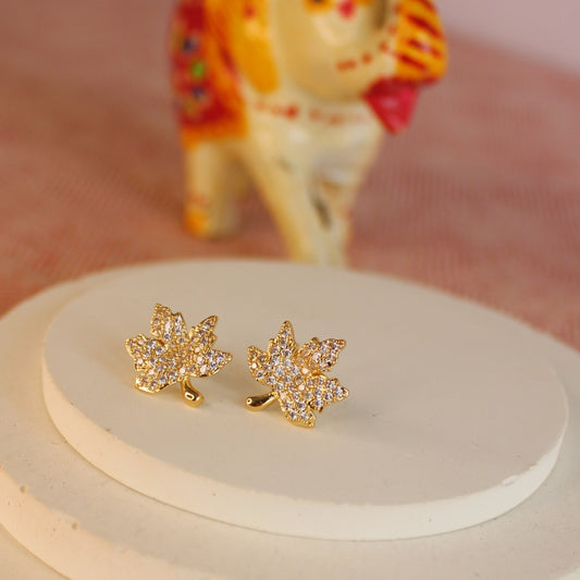 Gold leaf-shaped earrings with clear stones on a white stand with a colorful cat toy in the background.