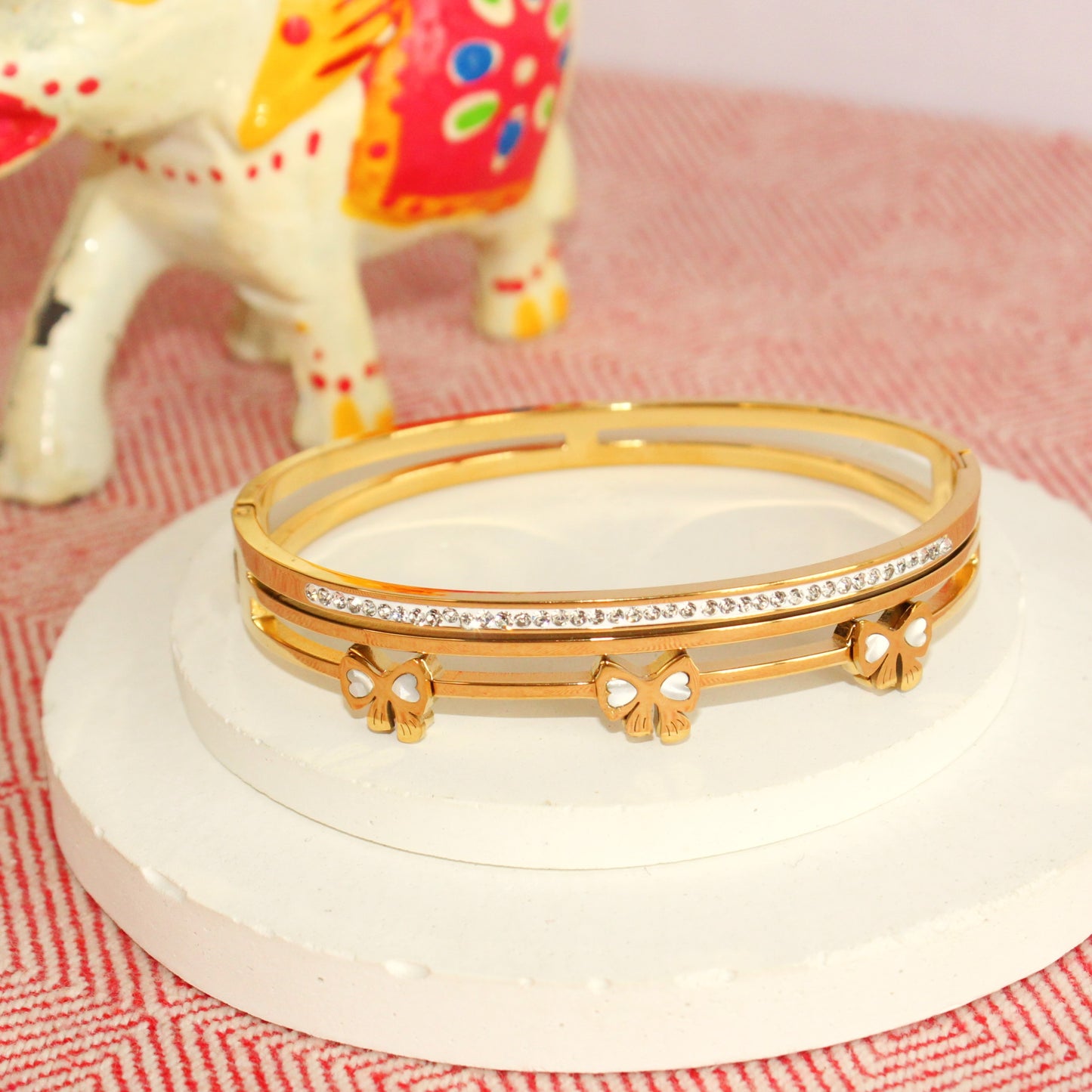 Gold bracelet with floral designs on a white stand with a colorful elephant figurine in the background.